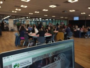 View over a laptop screen showing a room of people, (blurred), sitting around round tables talking. Stands and banners in the background.