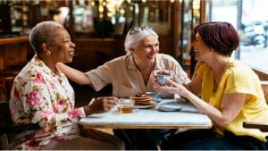 3 women sat at a cafe table talking and smiling. one has a coffee cup in her hand.